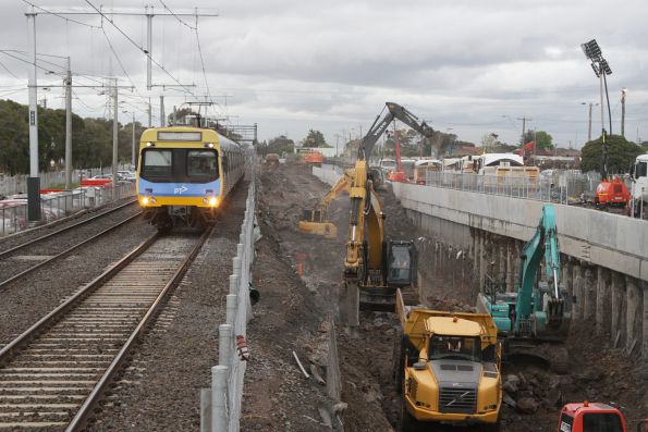 Down Comeng train approaches St Albans station, as excavators remove rock from the future rail cutting